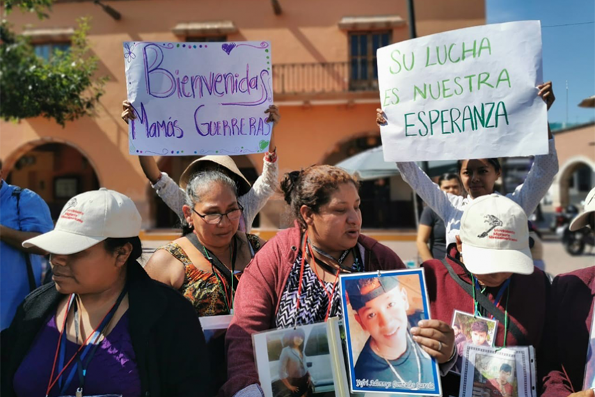 Women gather together outside holding signs that read 'Welcome mothers' and signs with photos of missing children