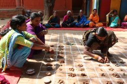 Women crouch outside over a grid drawn with chalk as they place seeds within the squares. The women are wearing colourful saris.