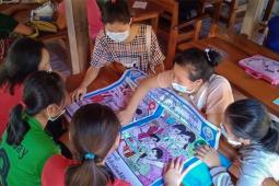 Six young women, each wearing a medical face mask, sit around a table looking at a poster of two situations: one marked with a red "X" and the other marked with a green check mark.