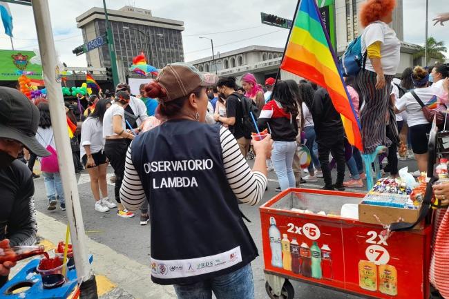 Photo du dos d'une personne portant un gilet avec l'inscription "obersvatorio lambda". Ils sont à la parade des fiertés et distribuent des friandises.