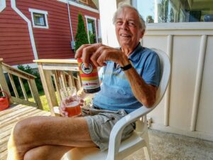 Photo of a smiling man sitting on a patio chair. He holds a beer