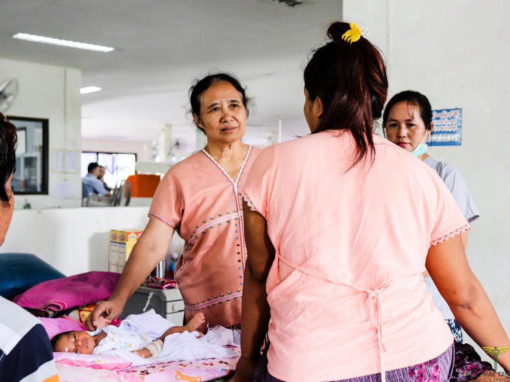 Woman in pink scrubs stands over a very young baby and looks at another woman wearing pink scrubs.