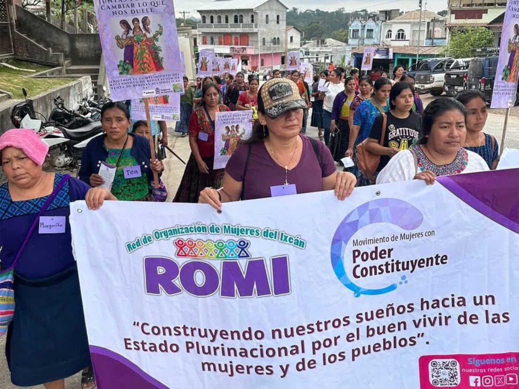A group marches down a street holding a large banner for ROMI and the Movimiento de Mujeres con Poder Constituyente, displaying the message about building a plurinational state for the well-being of women and communities. Behind the banner, participants carry illustrated signs. Buildings and vehicles line the background.