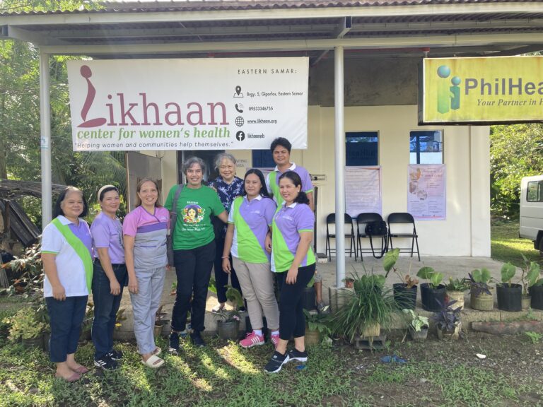 A group of people stands in front of a small clinic building with banners for Likhaan Center for Women’s Health and PhilHealth, surrounded by plants and outdoor chairs.