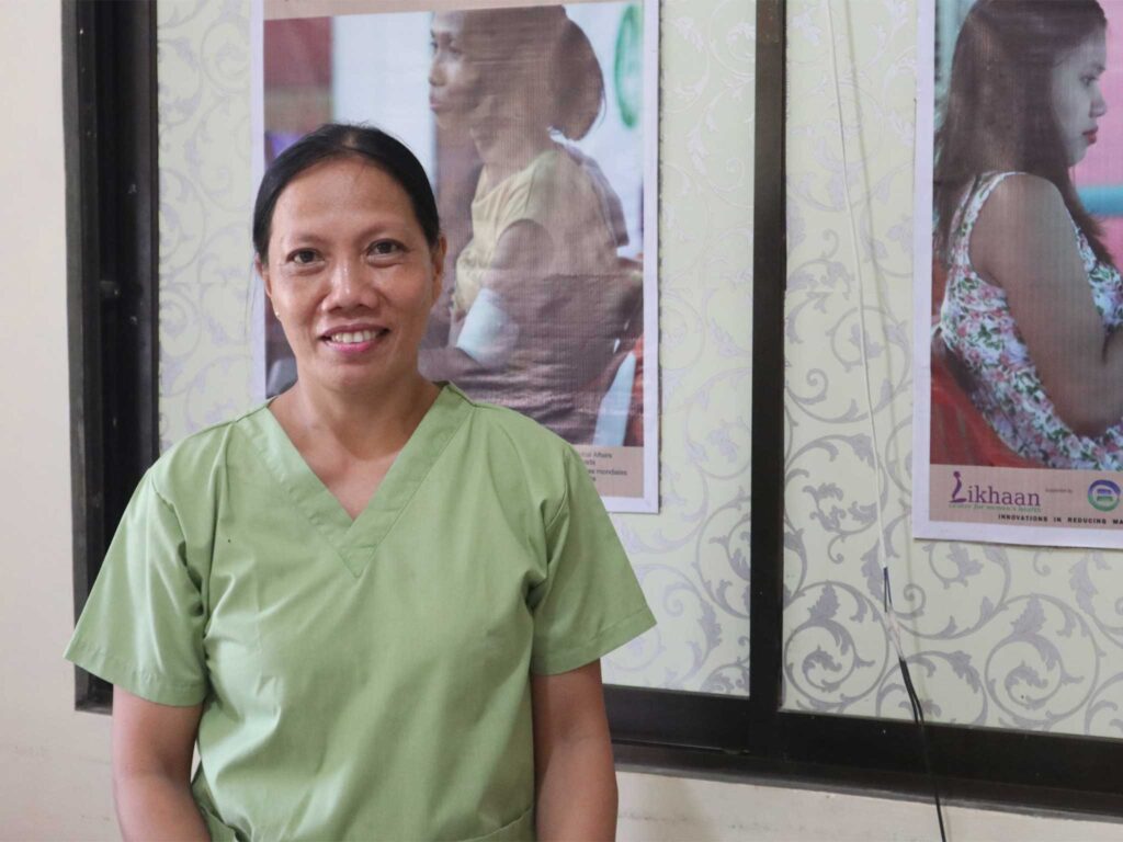 Melba wears a light green scrub-style top and stands in front of patterned wallpaper and two posters. The posters feature images related to family planning, including one showing a person holding a child and another showing a pregnant individual