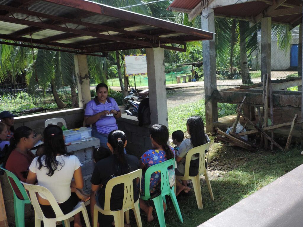 A group sits on plastic chairs under an open shelter while one person leads an educational session with materials on a table. Nearby, three others sit reading pamphlets in a rural outdoor setting with palm trees and simple wooden structures.