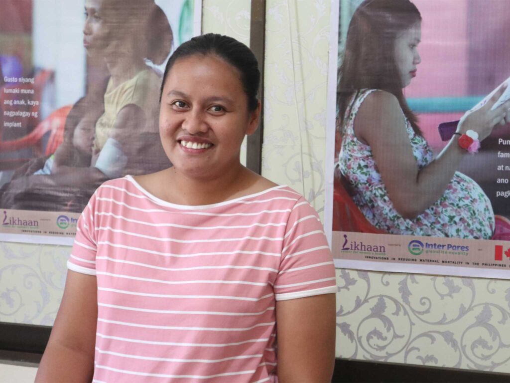 Jayza wears a pink and white striped shirt and sits in front of patterned wallpaper and two informational posters. One poster shows a person holding a baby, and the other shows a pregnant person reading a booklet.