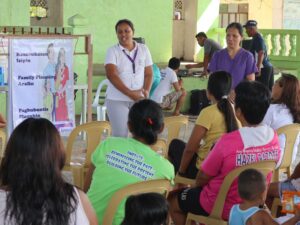 A community group sits on plastic chairs at an open-air venue while a facilitator leads a family planning session beside a poster display. Another staff member stands nearby as participants listen, with children and additional people visible.