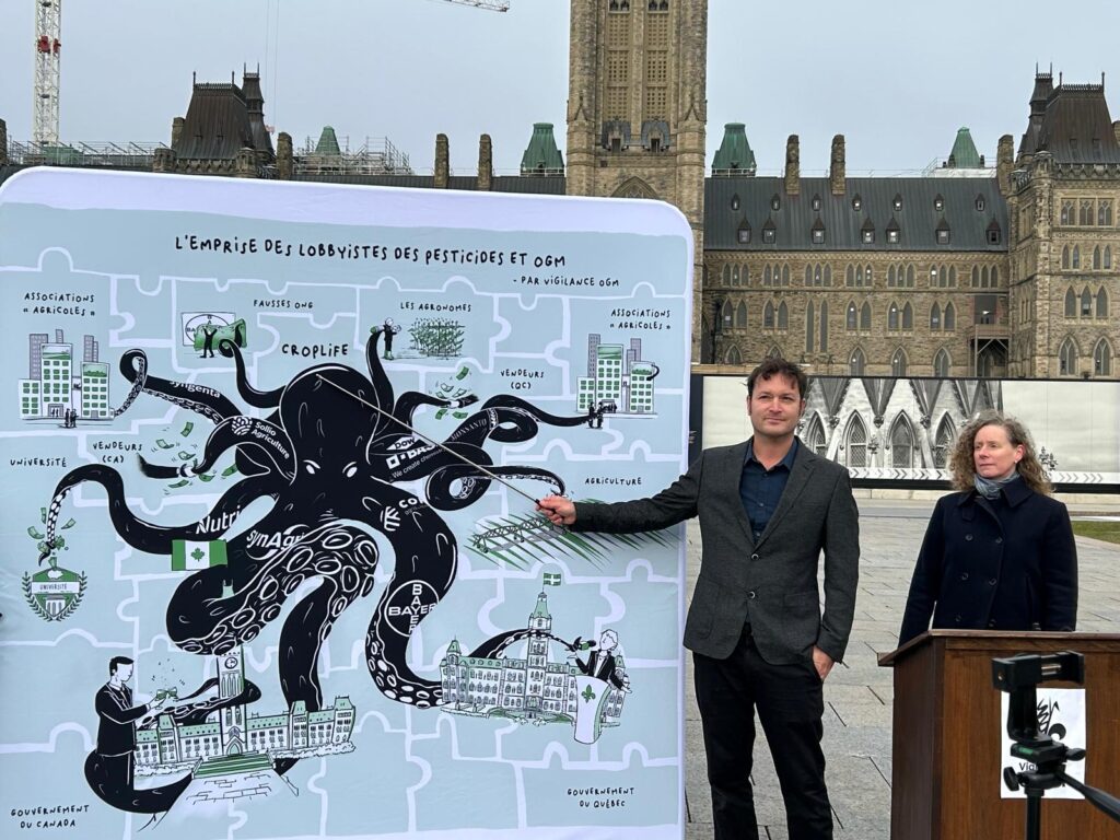 A man and a woman stand beside a large illustrated panel on Parliament Hill in Ottawa. The artwork depicts an octopus representing the influence of pesticide and GMO industry lobbyists, with tentacles reaching into government, universities, agricultural organizations, and other institutions.