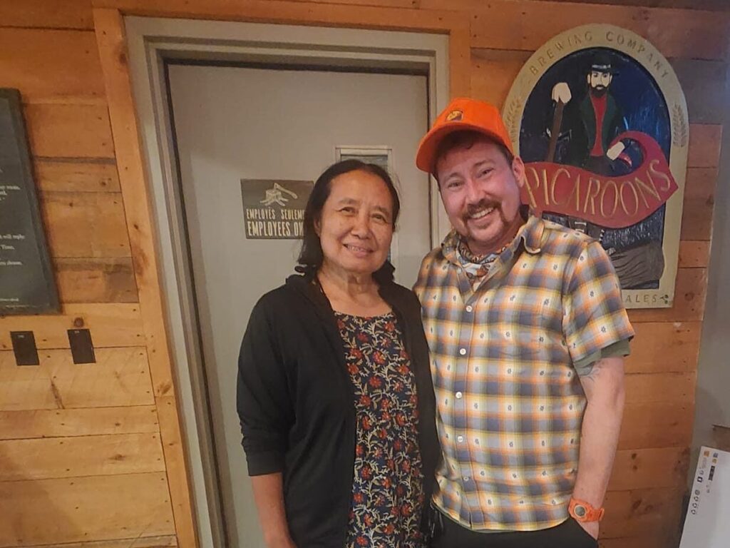 Two people pose for a photo inside a rustic, wood-paneled building. One person carries a handbag, while the other wears an orange cap and plaid shirt. A brewery sign is visible on the wall behind them.