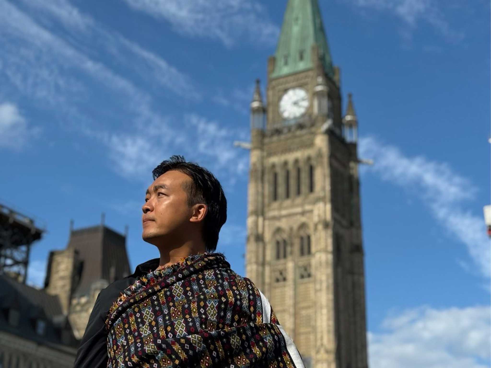 Une personne vêtue d'un tissu tissé aux couleurs vives se tient devant les bâtiments du Parlement canadien à Ottawa. La Tour de la Paix, avec son toit en cuivre vert caractéristique et son cadran d'horloge, se détache derrière elle sur fond de ciel bleu parsemé de nuages.