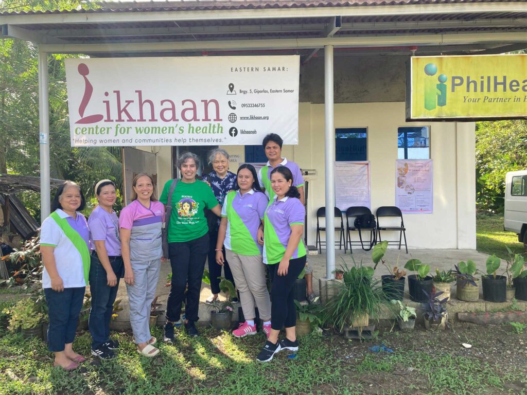 Un groupe se tient devant une petite clinique, sous l'enseigne du « Centre Likhaan pour la santé des femmes », dans la province de Samar-Est. Le porche est équipé de chaises et décoré d'affiches, tandis que des plantes en pot bordent la pelouse devant l'entrée.