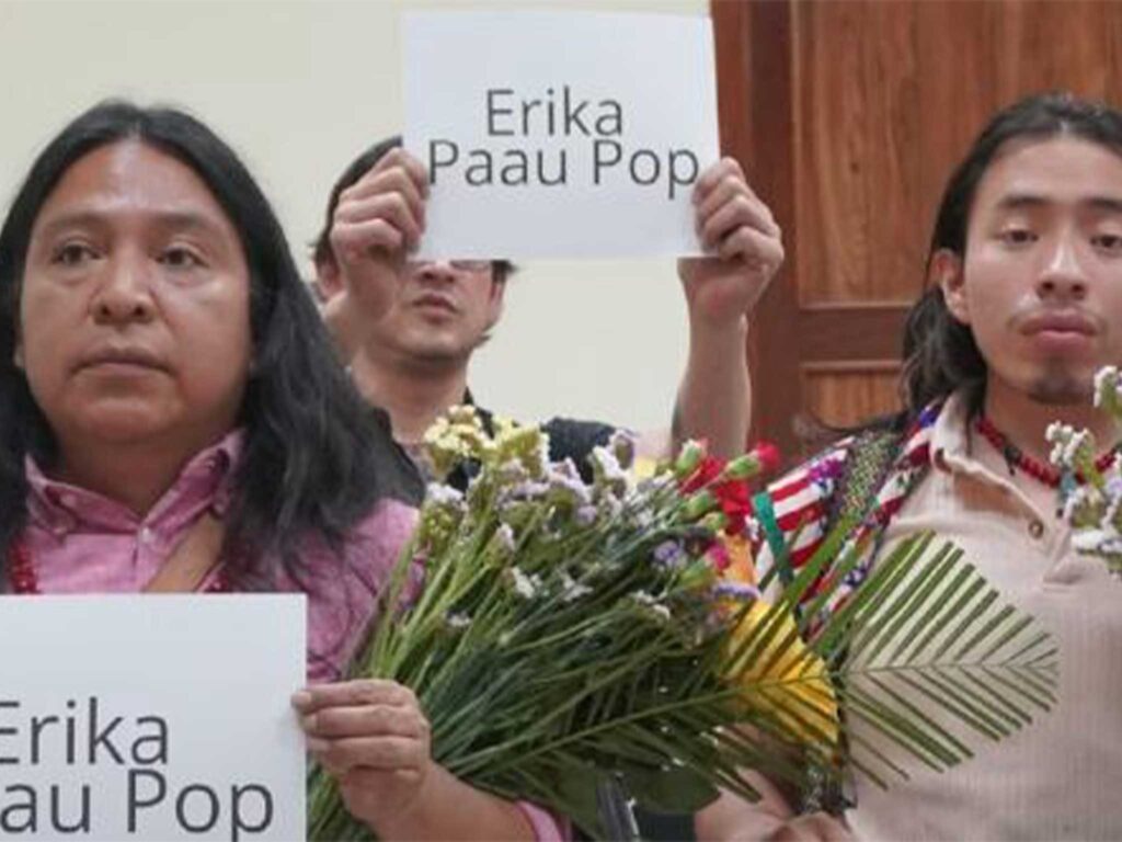 Three people holding signs reading Erika Paau Pop, standing indoors with flowers, positioned before wooden doors during a remembrance gathering.