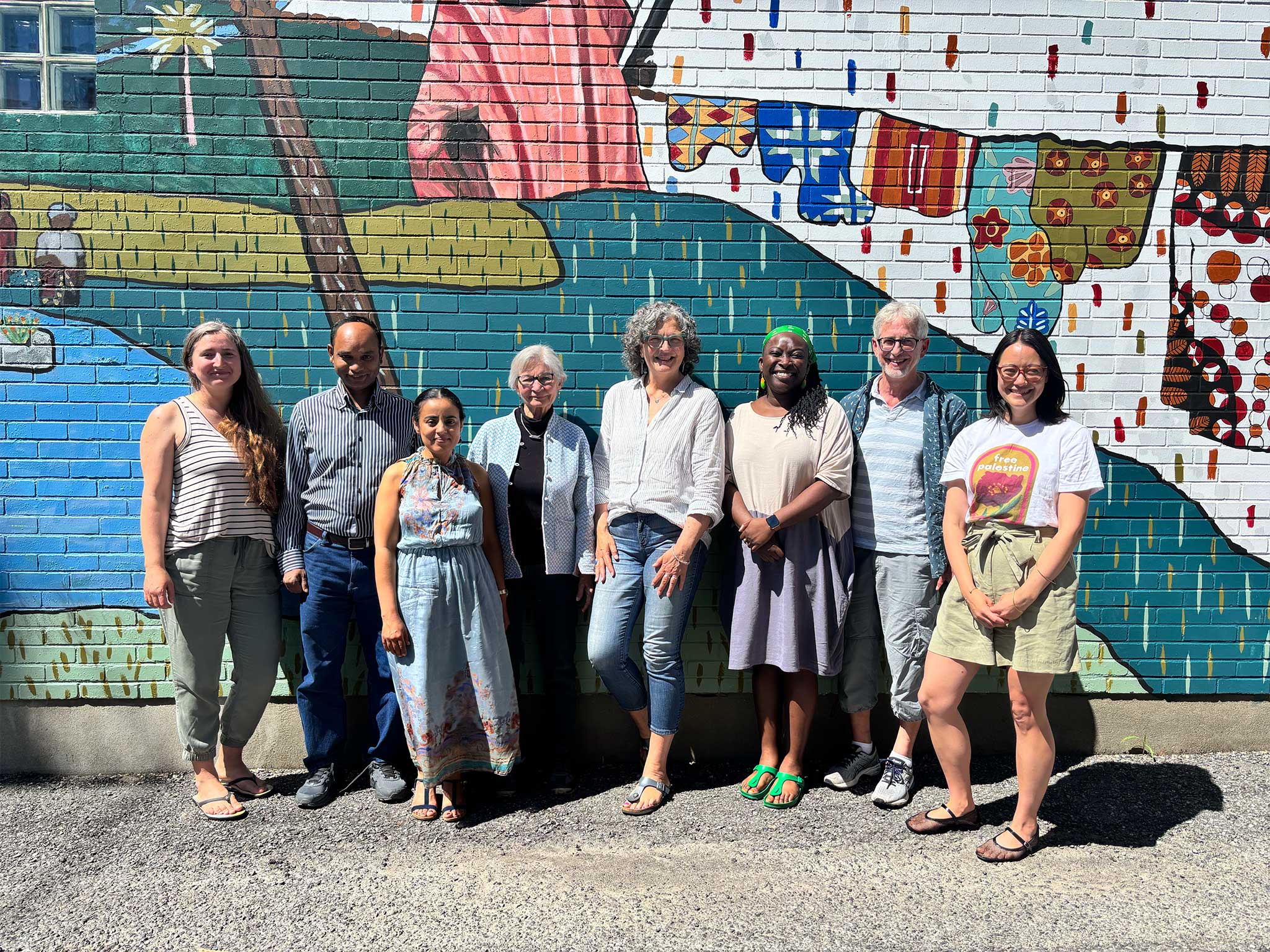A group of 8 people, Inter Pares Board members, stand together in front of a colourful mural. Everyone is smiling.