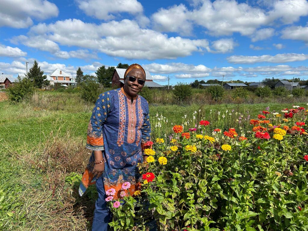 Mariamé Ouattara stands among vibrant flowers in a rural garden landscape beneath blue skies, celebrating agriculture, biodiversity and community.