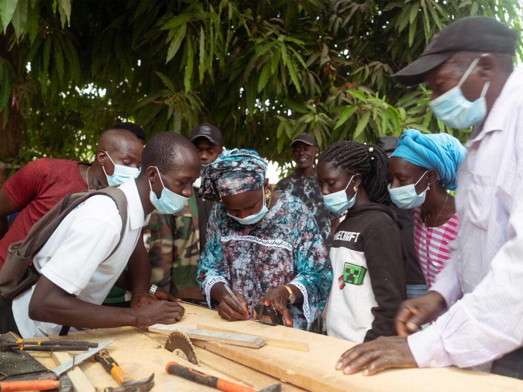 Community members gather around a workbench, learning woodworking techniques and measuring materials during practical skills training.