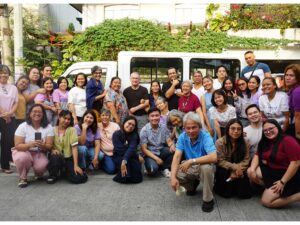 Large group of community members and staff gathered outdoors beside a white van, posing together in front of greenery.
