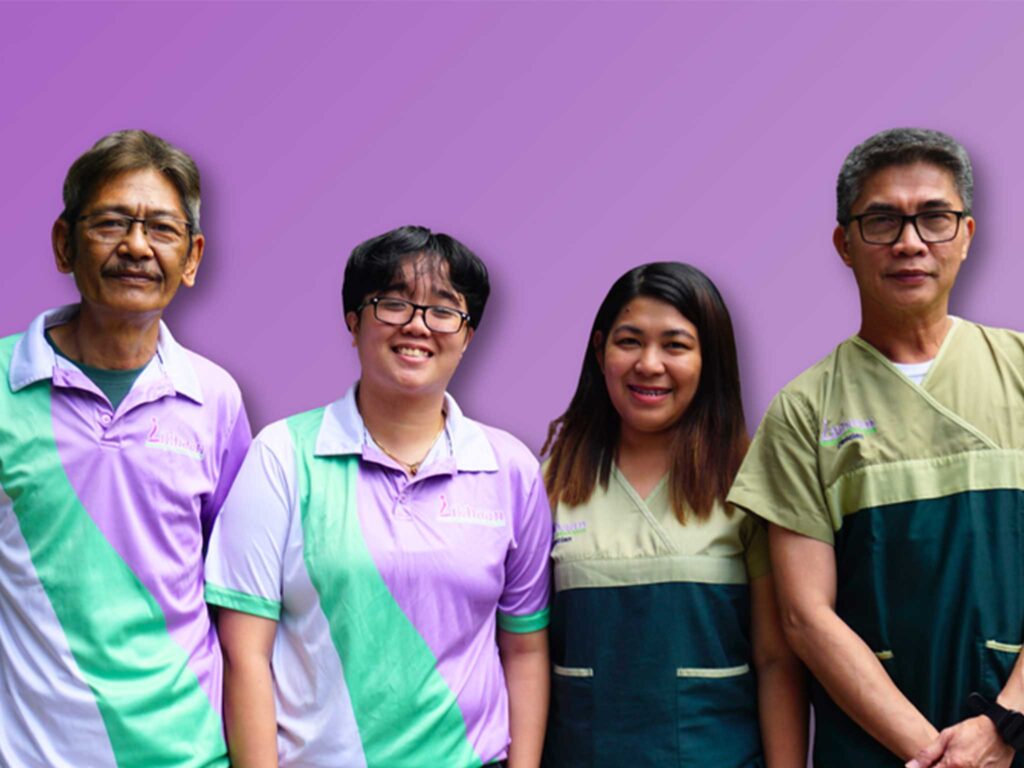 Four people stand side by side smiling at the camera. Two are wearing purple and green shirts. Two are wearing green and blue shirts.