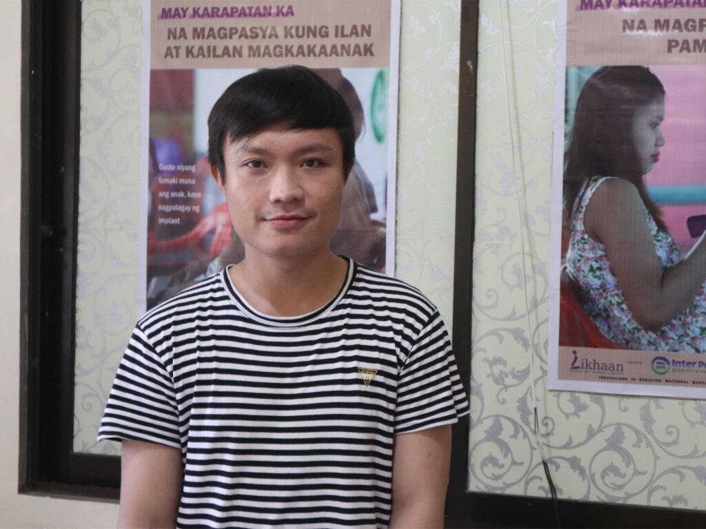 Michael Catipay wears a black and white striped t-shirt and stands in front of a poster detailing maternal health care.