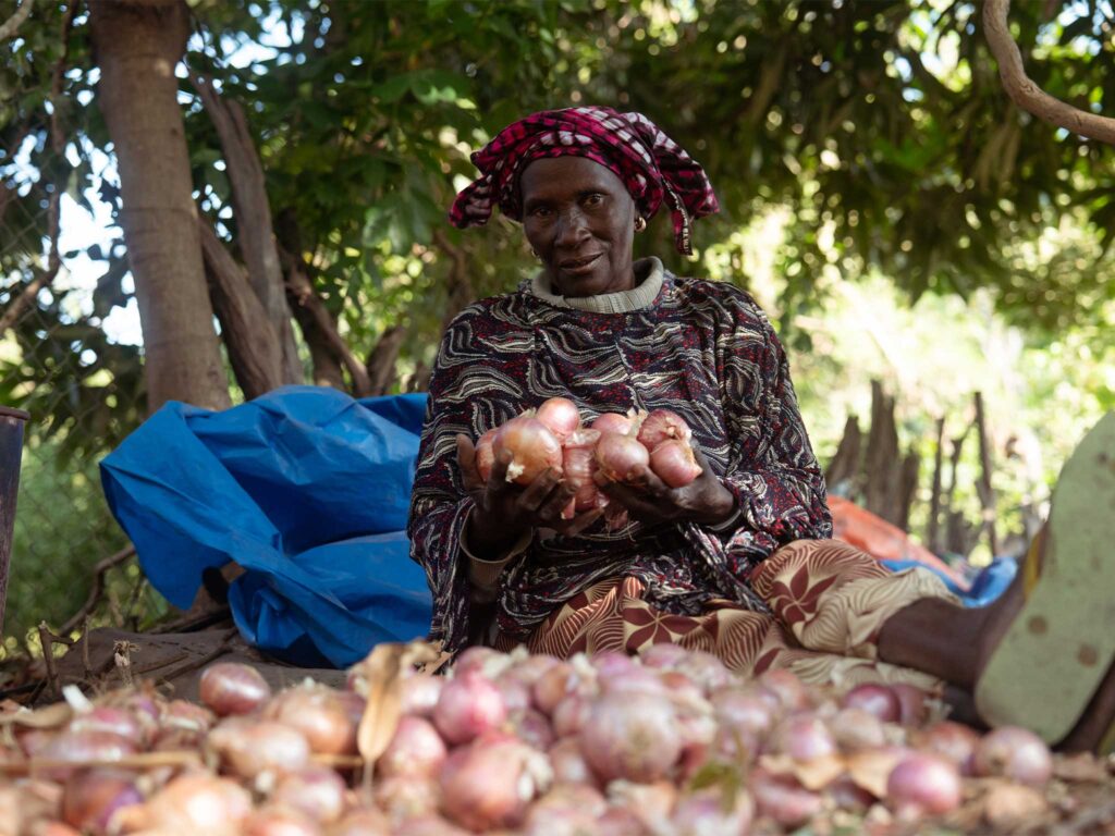 A woman farmer sits on the ground in front of a pile of onions, holding onions in her hands. She is smiling.